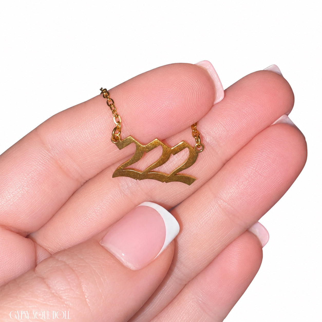 A gold-toned necklace with a stylised '222' pendant is held on a person's fingers against a plain white background.