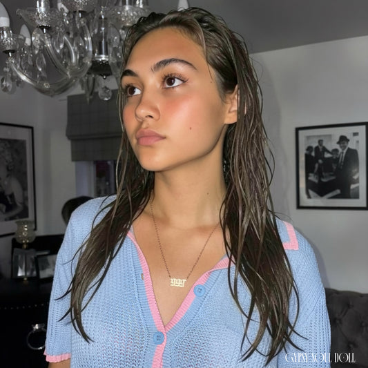 A model with wet-look hair wearing a blue and pink knit polo cardigan showcasing a gold 999 angel number nameplate necklace by Gypsy Soul Doll against a clean white backdrop.