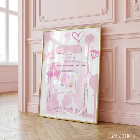 A framed art print featuring a pink Chanel Coco Mademoiselle perfume bottle illustration, leaning against a pink paneled wall on a light wood floor.