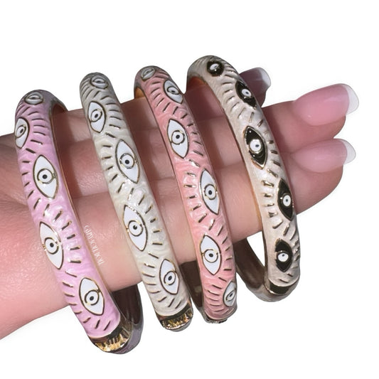 Close-up view of a hand with a classic french manicure showcasing a stacked collection of four enamel evil eye bangles in contrasting pastel pink and creamy white shades, detailed with gold accents and repeating protective eye motifs.