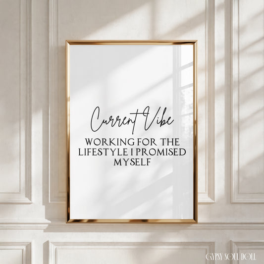 A framed typographic wall art print with a gold frame hangs on a white paneled wall. The print has a white background with black text reading 'Current Vibe' and 'WORKING FOR THE LIFESTYLE I PROMISED MYSELF.