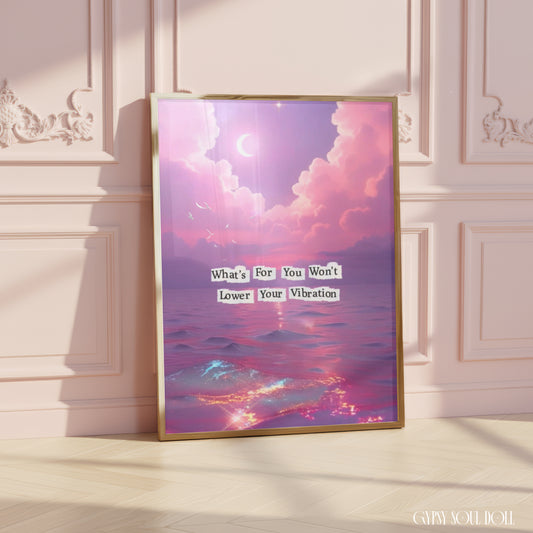 A framed art print leaning against a pink paneled wall on a light wood floor. The print features a pastel purple and pink sky with clouds, a crescent moon, and ocean waves, with the text 'What's For You Won't Lower Your Vibration'.