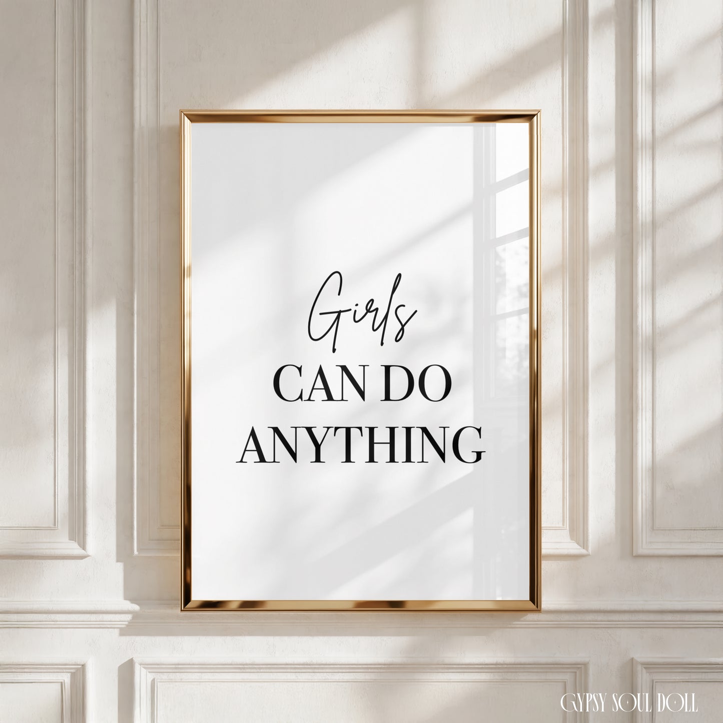 A framed typographic wall art print with the text 'Girls CAN DO ANYTHING' in black lettering on a white background, displayed against a white paneled wall with soft window shadows.