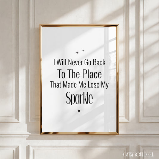 A framed typographic wall art print with the quote 'I Will Never Go Back To The Place That Made Me Lose My Sparkle' in black text on a white background, set in a gold frame and mounted on a white panelled wall with soft window shadows by Gypsy Soul Doll.
