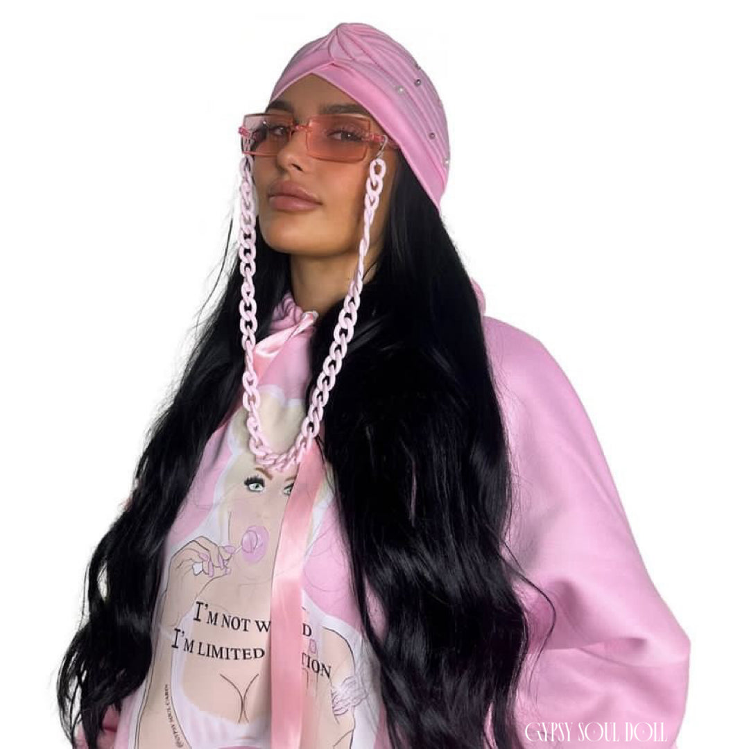 Close-up of a woman wearing a pink hoodie with a graphic of a woman holding a lollipop and the text 'I'm not weird, I'm limited edition,' accessorised with pink fashion sunglasses, and a matching pink bohemian gypsy headscarf, against a white background.