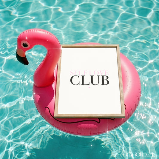 A pink flamingo-shaped pool float floating in a swimming pool, holding a square framed print with the words 'SELF LOVE CLUB' on a white background.
