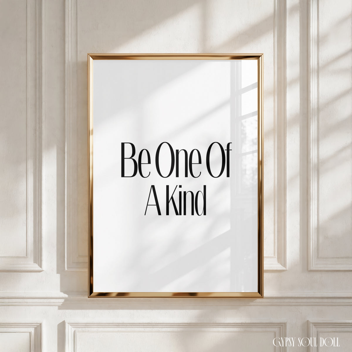 A framed typographic wall art print with the phrase 'Be One Of A Kind' in black serif text on a white background, set in a thin gold frame against a cream panelled wall with soft window shadows.