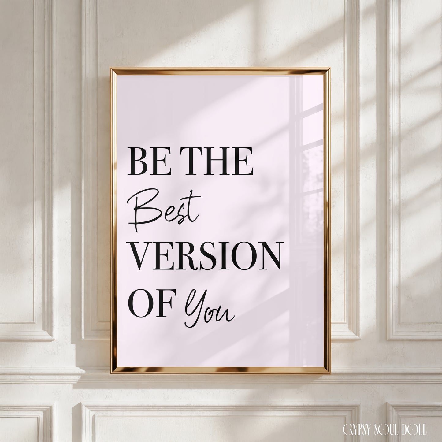 A framed typographic wall art print with the text 'BE THE Best VERSION OF You' in black lettering on a pale pink background, displayed against a white panelled wall with soft window shadows.