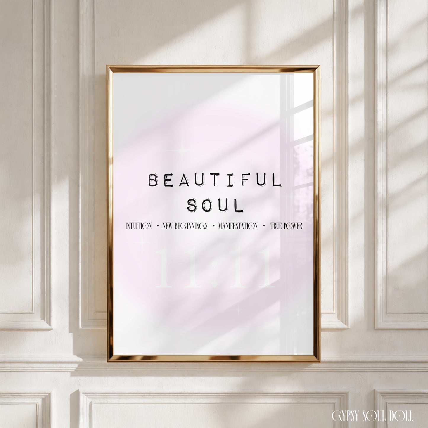 A framed art print with the text 'BEAUTIFUL SOUL' and smaller angel number manifestation meanings, displayed on a white panelled wall with soft window shadows. The print has a white background with a faint pink glow and is enclosed in a thin gold frame.