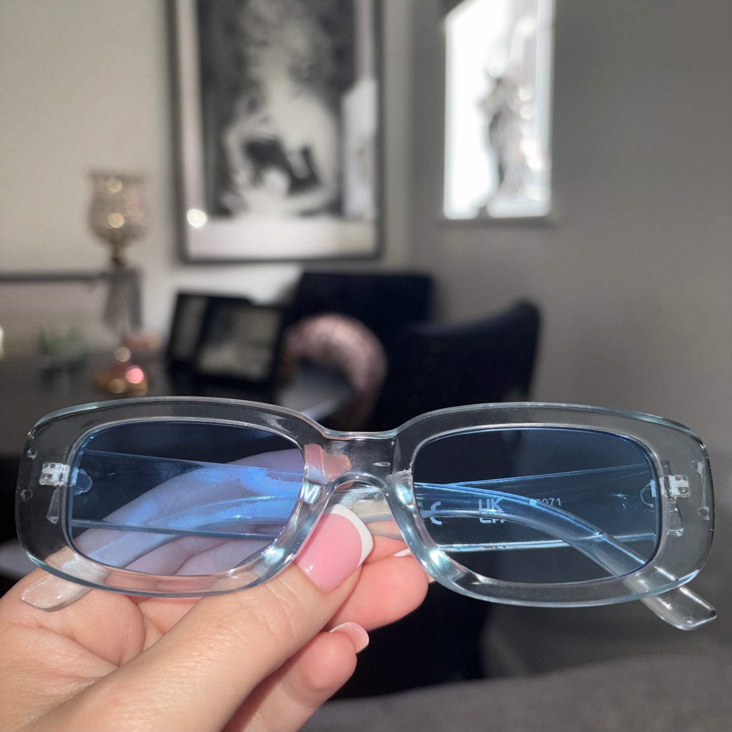 A person's hand holding a pair of translucent light blue square sunglasses with blue-tinted lenses in a modern, blurred living room setting.