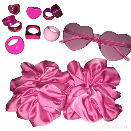 A curated Y2K candy pink accessory bundle featuring two oversized pink satin scrunchies, a pair of frameless translucent heart-shaped fashion glasses, and an assortment of retro resin rings including 3D puffy hearts, metallic pink bands, and chunky acrylic domes on a plain white background.