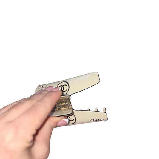 A hand holding a cream-coloured hair claw clip with a gold-toned metallic center and black printed Chanel logo details, set against a plain white background.