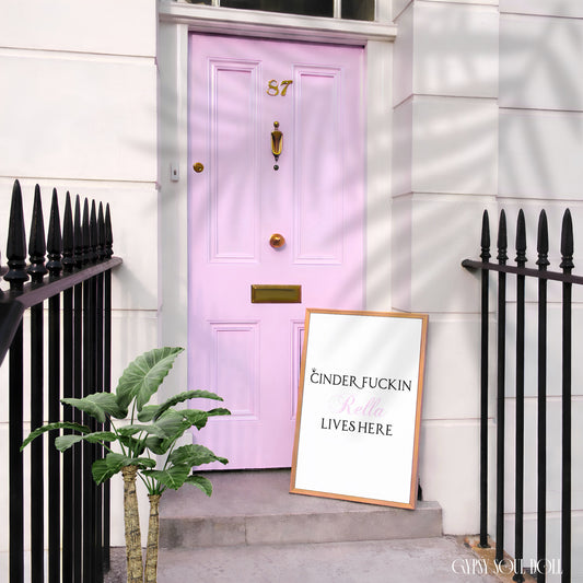 A framed art print with the text 'CINDER, FUCKIN' DONT LET THEM LIVES HERE' in black and pink lettering on a white background, leaning on the steps next to a light pink front door.