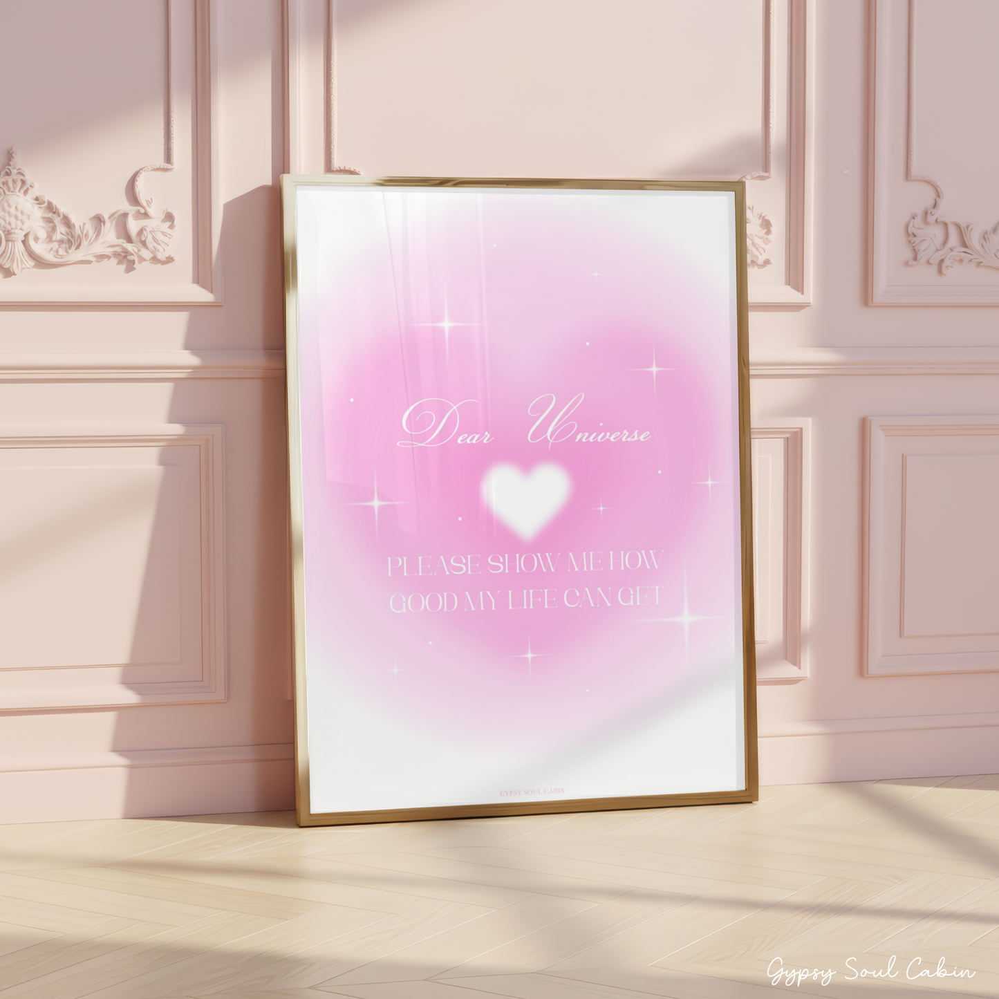 Minimalist A4 manifestation wall art print featuring the text Dear Universe in elegant script and PLEASE SHOW ME HOW GOOD MY LIFE CAN GET in a clean white serif font over a dreamy pink aura heart gradient background with sparkling stars, leaning in a gold frame on a wooden floor against a pink panelled wall.