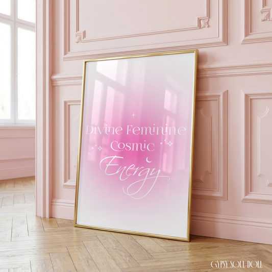Minimalist A4 celestial manifestation wall art print featuring the text Divine Feminine Cosmic Energy in an elegant white script and serif layout over a soft pink aura gradient backdrop with line-art stars, leaning in a gold frame on a wooden floor against a pink panelled wall.