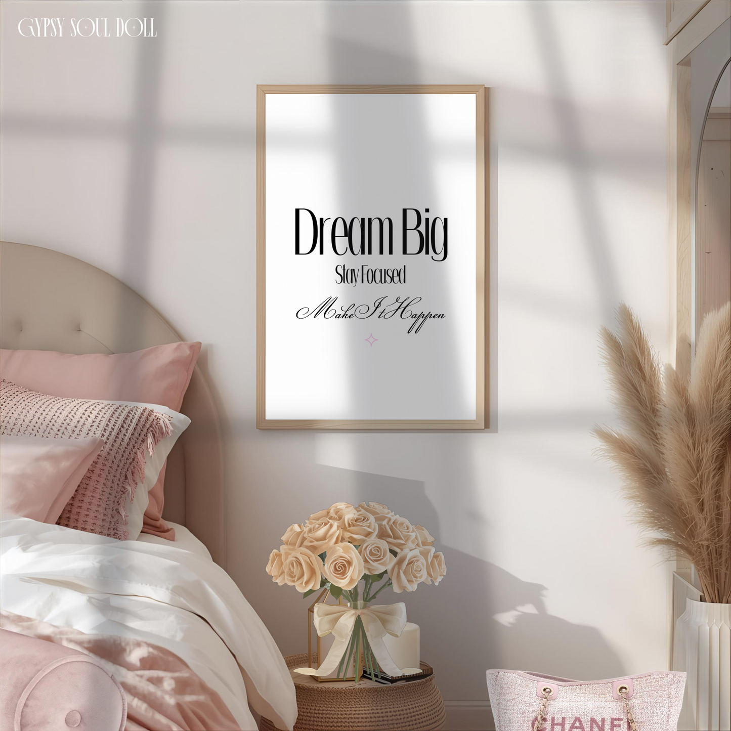 Minimalist motivation typography print reading Dream Big Stay Focused Make It Happen in a light wood frame hanging above a cosy bed with pink pillows and a bedside bouquet of cream roses.