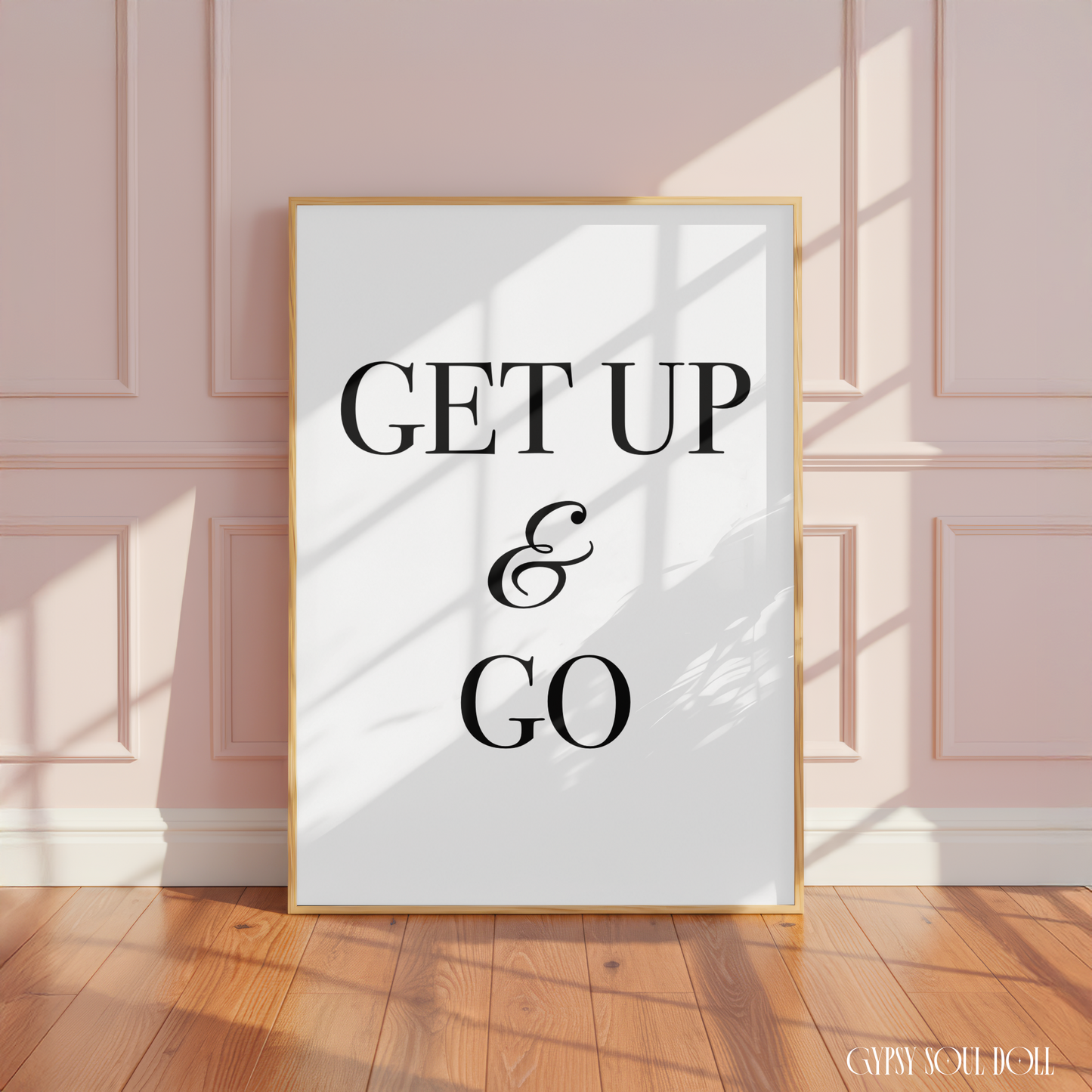 Minimalist black and white manifestation affirmation typography print reading Get Up & Go with a cursive ampersand in a large oak frame leaning against a panelled pink wall.