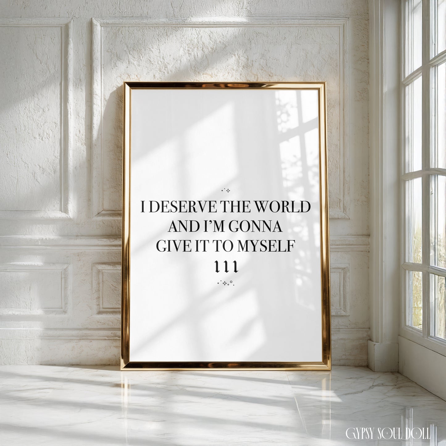 A vertical framed typographic art print with a gold frame leans against a white paneled wall in a sunlit room. The print has a white background with black serif text reading, 'I DESERVE THE WORLD AND I'M GONNA GIVE IT TO MYSELF 111.