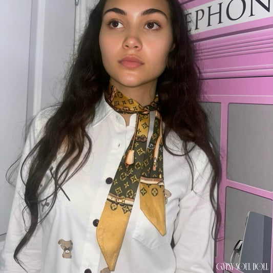 Model showcasing the Gypsy Soul Doll khaki and gold monogram skinny satin scarf wrapped elegantly as a multi-way neck tie choker, styled next to a pink telephone box.
