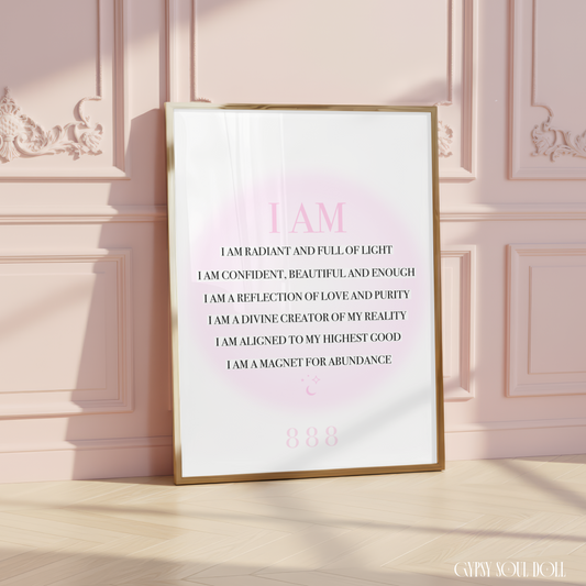 A framed inspirational quote print leaning against a pink paneled wall on a light wood floor. The print has a gold frame, a white background, and pink text that reads, 'I AM I AM RADIANT AND FULL OF LIGHT I AM CONFIDENT, BEAUTIFUL AND ENOUGH I AM A REFLECTION OF LOVE AND PURITY I AM A DIVINE CREATOR OF MY REALITY I AM ALIGNED TO MY HIGHEST GOOD I AM A MAGNET FOR ABUNDANCE.