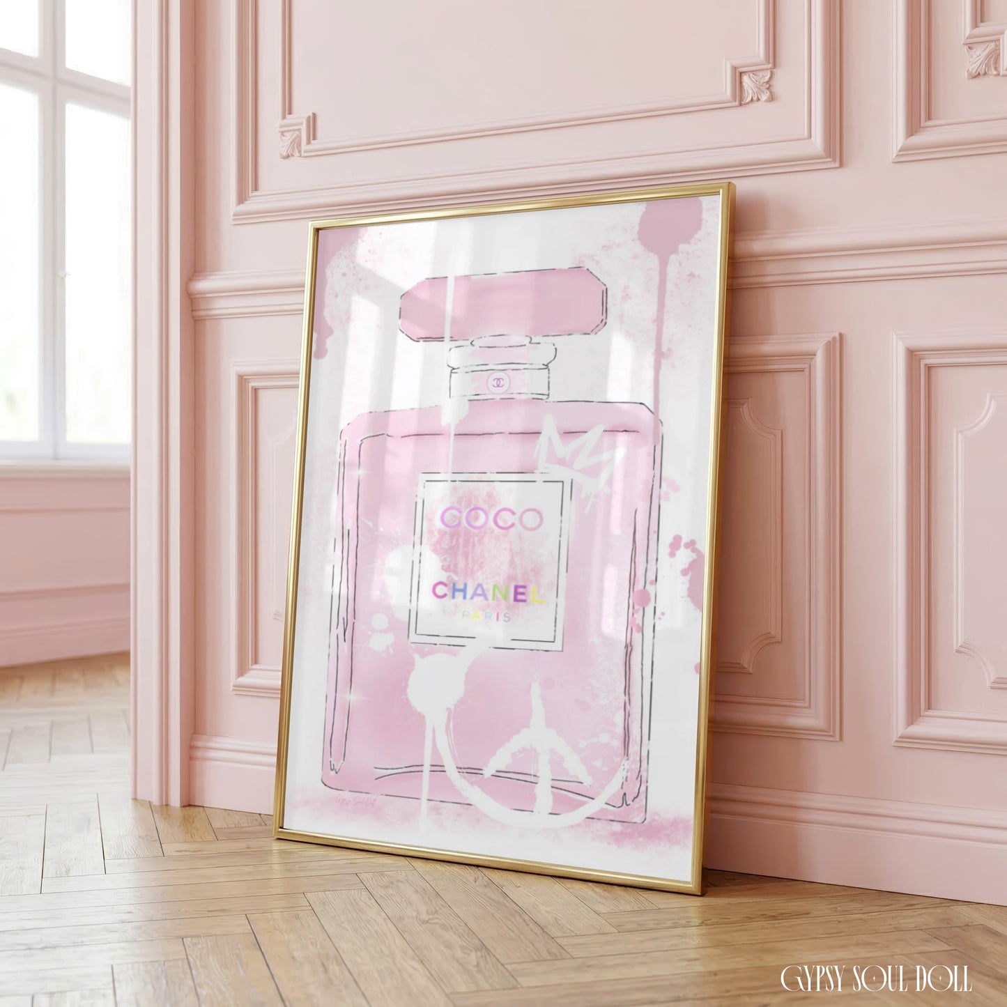 A framed art print featuring a pink Chanel Coco perfume bottle illustration, leaning against a pink paneled wall on a light wood floor.