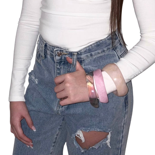 Lifestyle view of a model wearing the three-piece pink and iridescent acrylic bangle stack on her wrist, paired with distressed blue jeans and a white long-sleeve top.