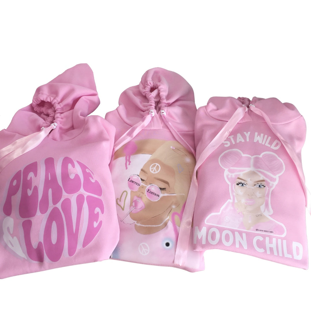 Flat lay of three pink hoodies including the Peace Love graphic, Spiritual Girl design, and Stay Wild Moon Child art with custom satin ribbon drawstrings.