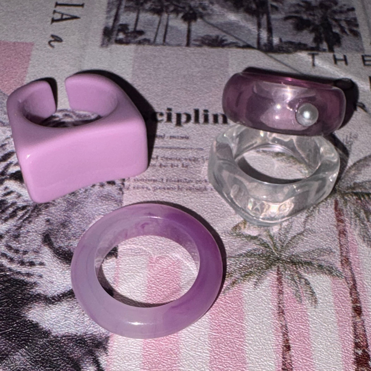 Four rings displayed on a patterned fabric surface: a chunky lavender ring, a clear faceted ring with a purple top and pearl detail, and a smooth lavender ring.