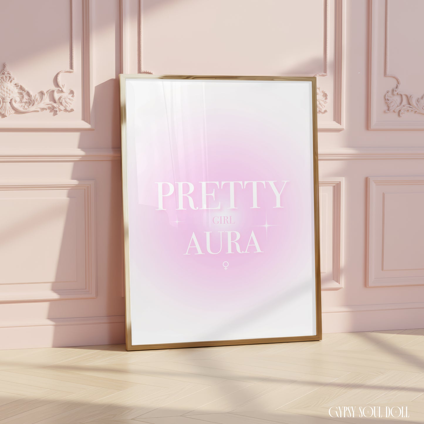 A framed art print with a gold frame leans against a pink paneled wall on a light wood floor. The print features a soft pink and white gradient background with the text 'PRETTY GIRL AURA' in white.