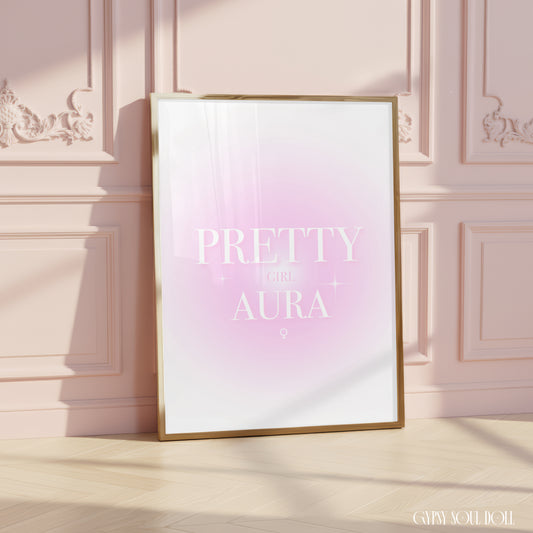 A framed art print with a gold frame leans against a pink paneled wall on a light wood floor. The print features a soft pink and white gradient background with the text 'PRETTY GIRL AURA' in white.