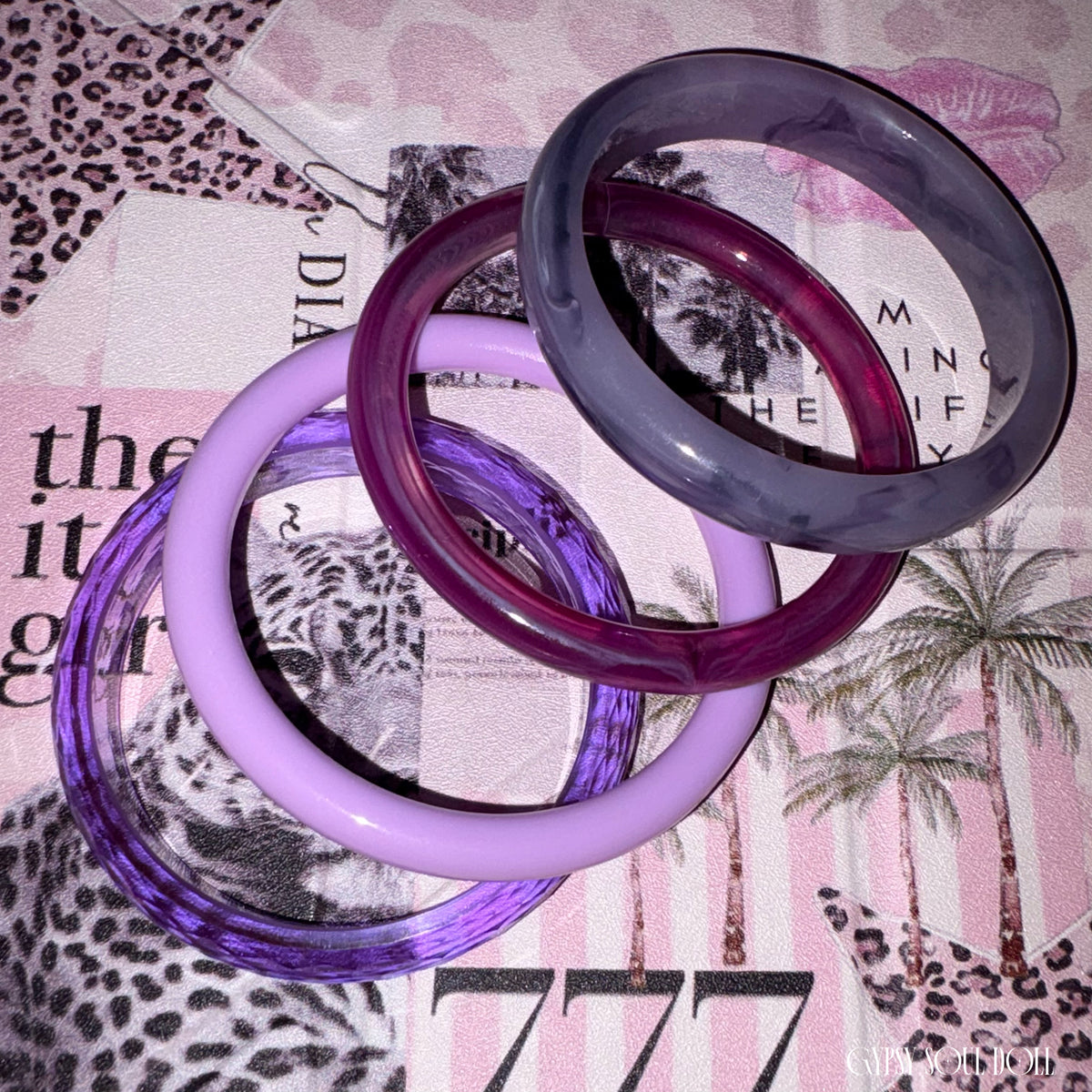 Four stacked translucent bangles in different shades of purple and lavender, photographed on a patterned pink and white surface by Gypsy Soul Doll.
