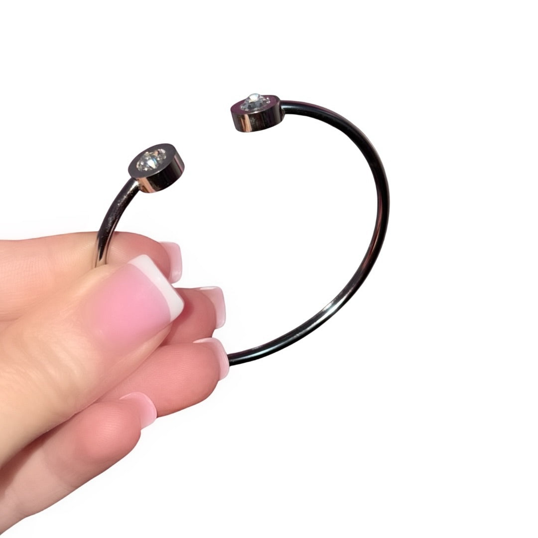 Side profile view of a fingers with a soft pink French manicure holding a polished silver open cuff bangle bracelet featuring two circular ends inset with bright cubic zirconia stones and engraved Roman numerals.