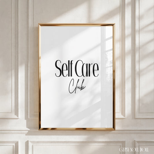 A framed wall art print with the words 'Self Care Club' in black lettering on a white background, set in a gold frame and mounted on a white panelled wall with soft window shadows by Gypsy Soul Doll.