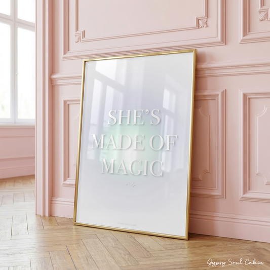 Minimalist A4 celestial print featuring the text SHE'S MADE OF MAGIC in bold white typography over a soft pastel green aura gradient background, leaning in a gold frame on a wooden floor against a pink panelled wall.