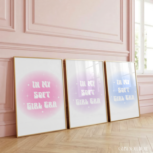 Three In My Soft Girl Era aura gradient typography prints in pink, purple, and blue, styled in luxury gold frames, leaning against a soft pink panelled studio wall on herringbone wood flooring.