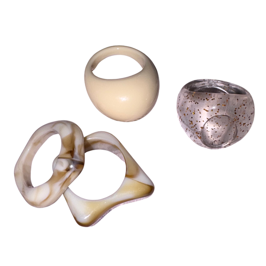 Four sculptural resin rings displayed on a solid white background. The set includes a chunky cream-colored ring with a loop, a smooth cream-colored bowl-shaped ring, and a translucent speckled brown ring.