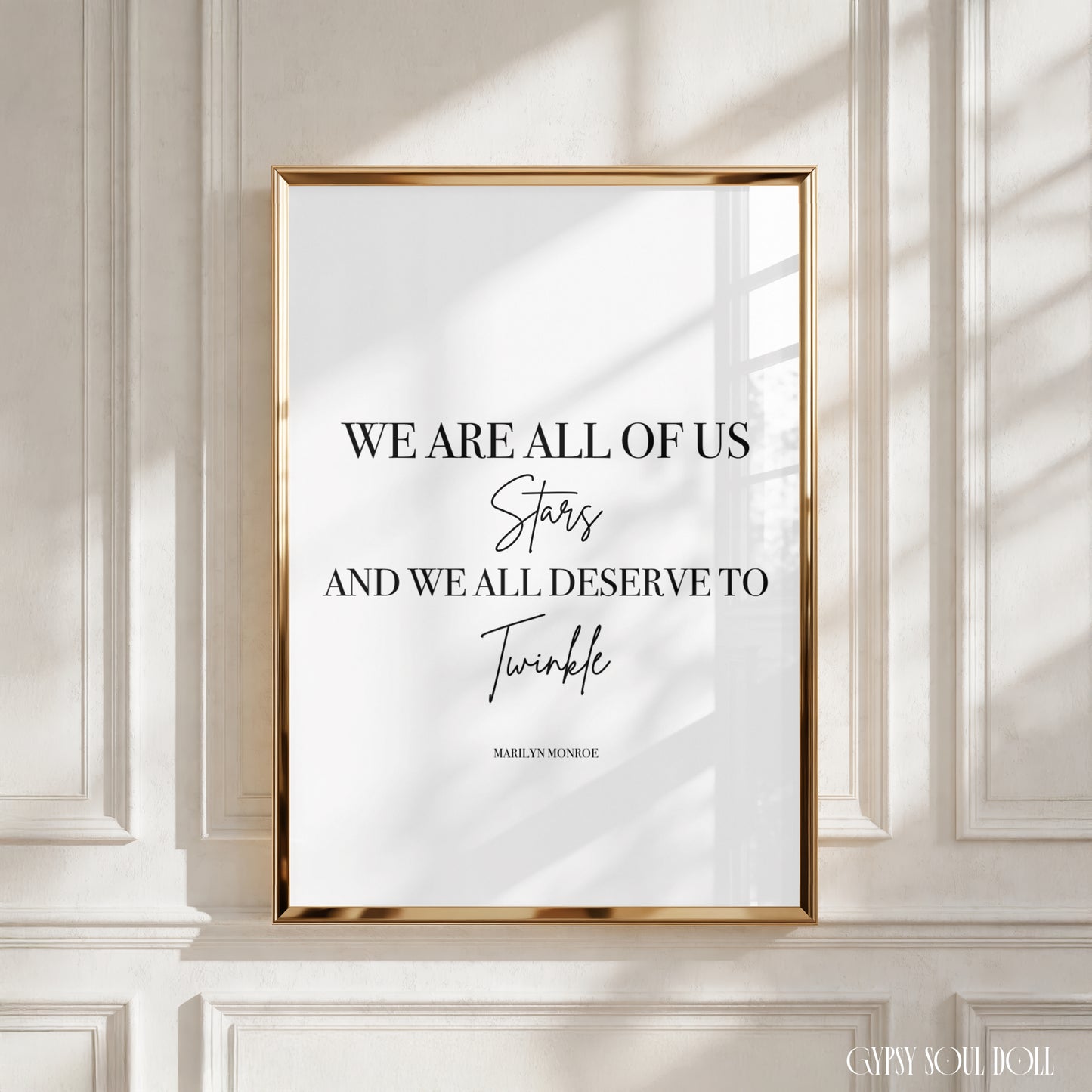 A framed typographic wall art print with a gold frame hangs on a white panelled wall. The print features black text reading, 'WE ARE ALL OF US Stars AND WE ALL DESERVE TO Twinkle' with 'Marilyn Monroe' printed below, and soft window shadows fall across the artwork and wall.