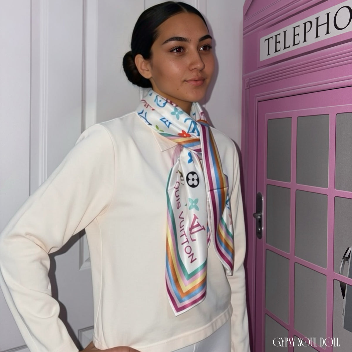 A model wearing a cream-coloured loungewear set and a colourful patterned silk scarf, standing next to a pink telephone booth.