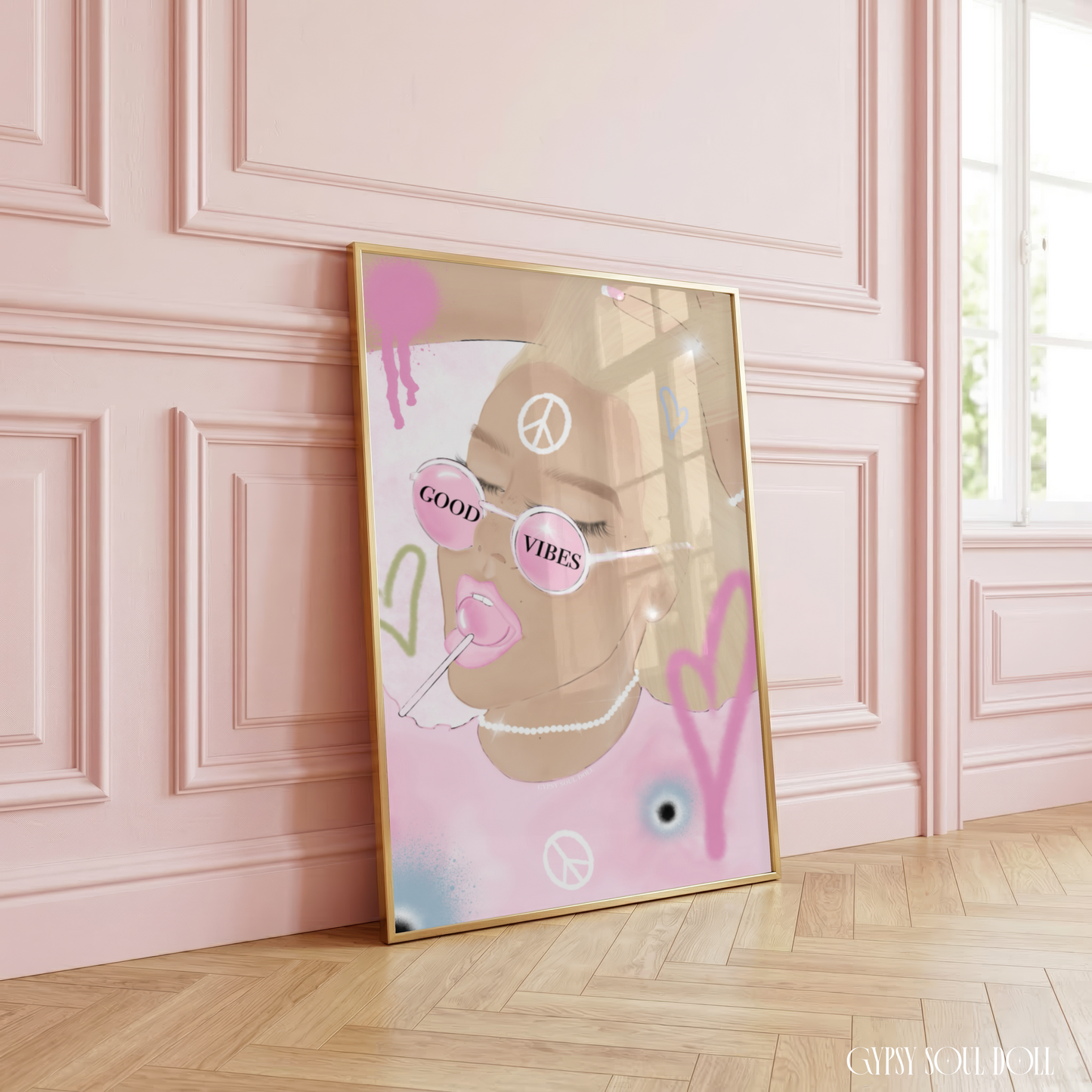 Trendy A4 aesthetic fashion print illustration of a girl with pink round sunglasses reading GOOD VIBES, eating a lollipop, decorated with graffiti spray paint hearts, peace symbols, and paint splatters, leaning in a gold frame on a wooden floor against a pink panelled wall.