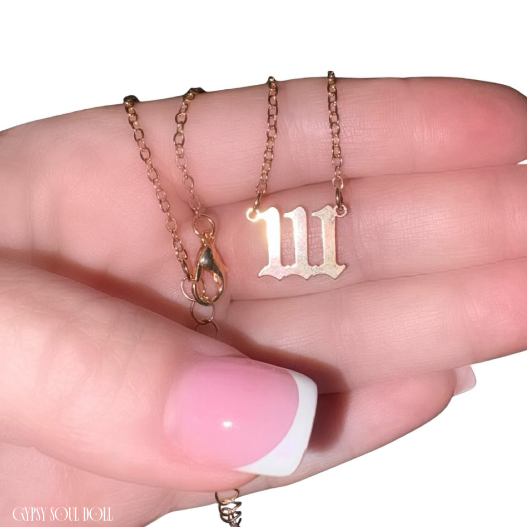 A close-up of a hand holding a delicate 111 angel number gold chain necklace with a Gothic-style letter 'W' pendant, against a clean white background.