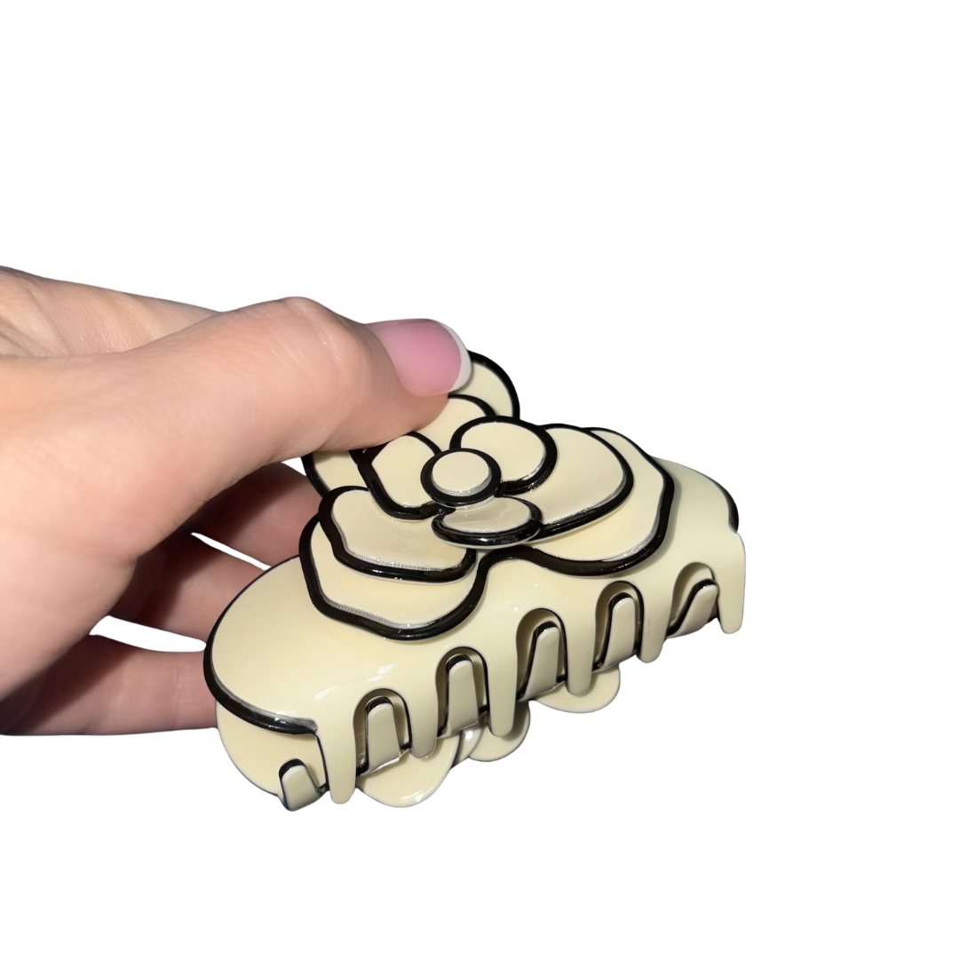 A hand holding a glossy ivory camellia flower hair claw clip with detailed black contrast outlines.