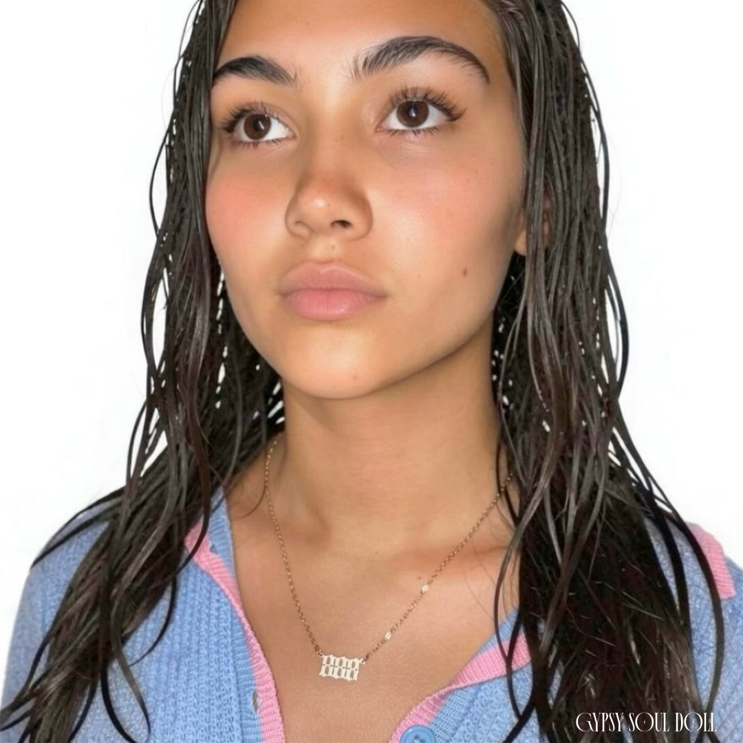 A close-up portrait of a model with wet dark hair wearing a delicate gold chain necklace with an 'angel numbers 888' pendant set against a plain white background.