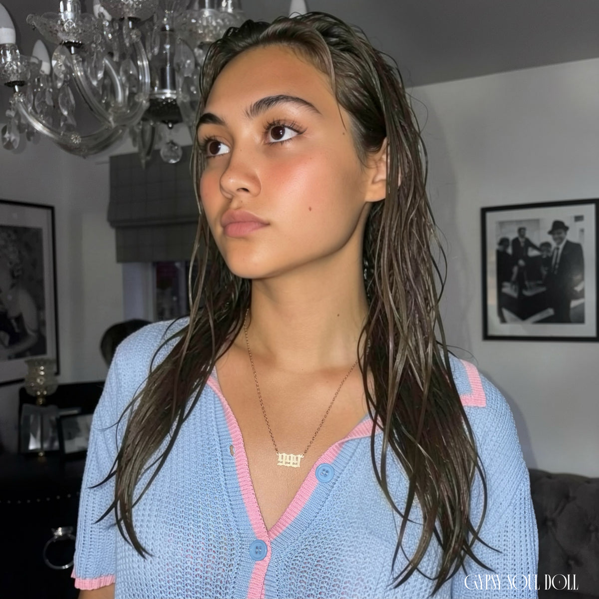 A model with wet-look hair wearing a blue and pink knit polo cardigan showcasing a gold 999 angel number nameplate necklace by Gypsy Soul Doll against a clean white backdrop.