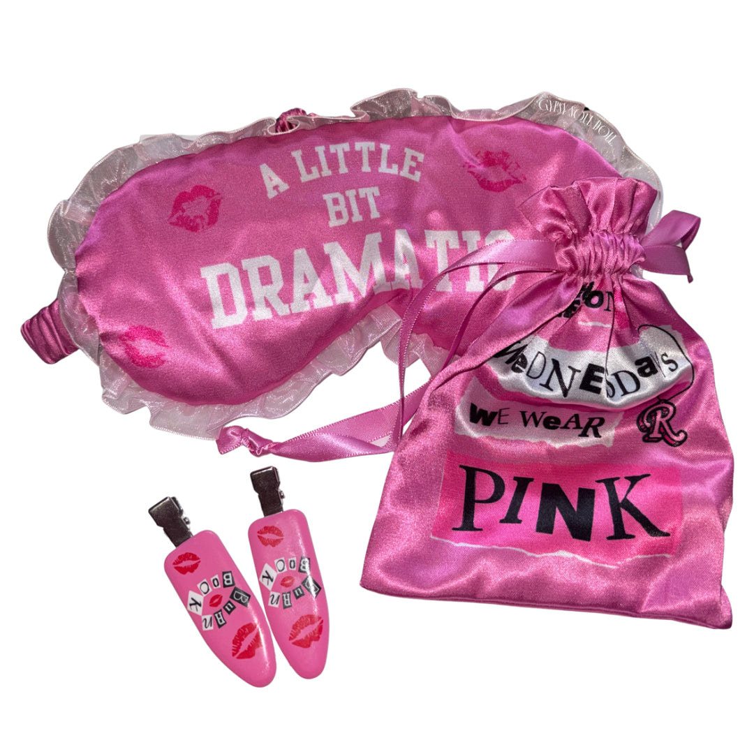 Flat lay view of a hot pink sleep set featuring an eye mask printed with A Little Bit Dramatic, a matching satin drawstring pouch reading On Wednesdays We Wear Pink, and two pink hair clips decorated with Burn Book lettering and red kiss marks.