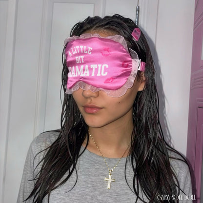 Side profile view of a girl with long damp dark hair wearing a hot pink satin eye mask featuring the text A Little Bit Dramatic with vibrant kiss marks and white ruffle trim, standing in front of a pink telephone booth door by Gypsy Soul Doll.
