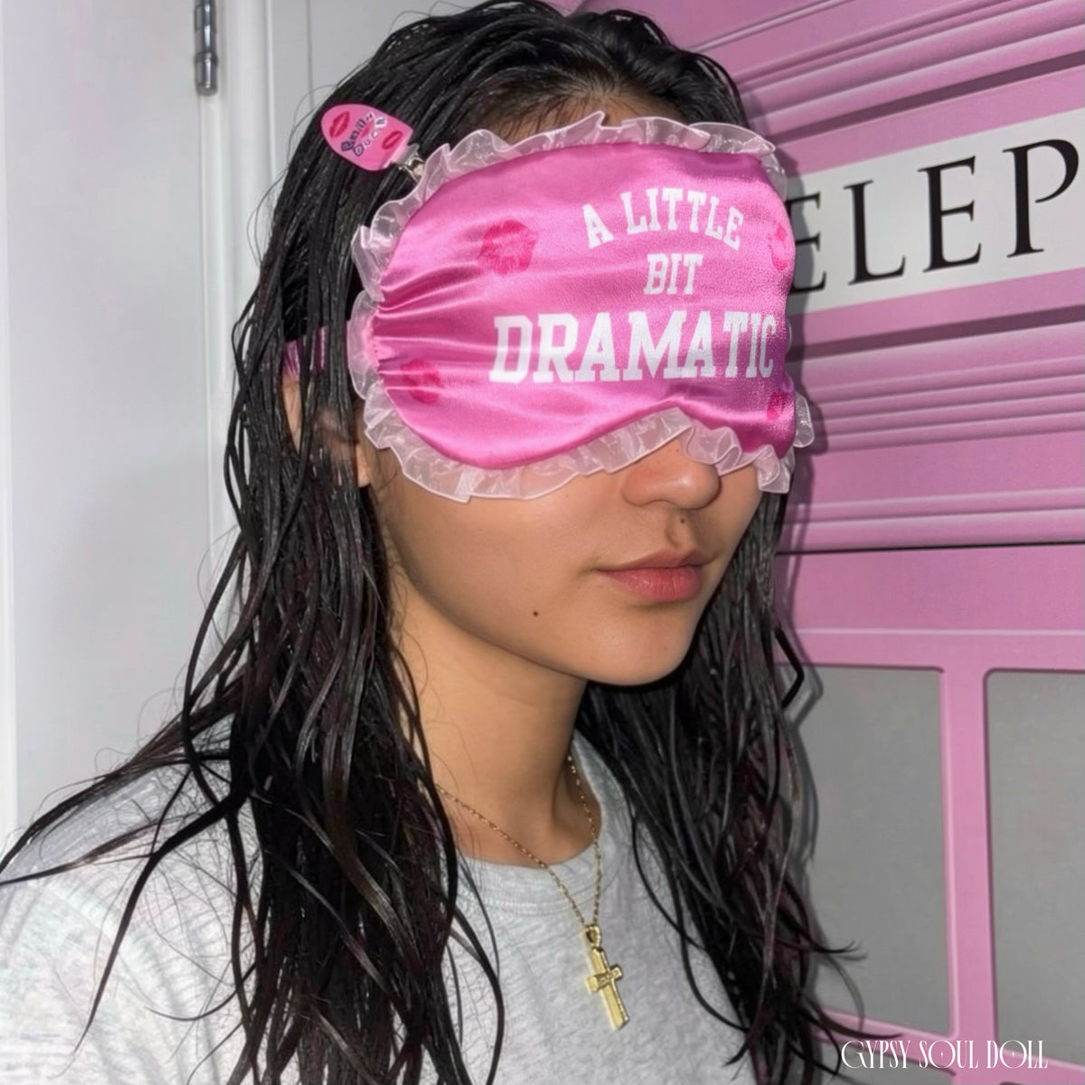 Side profile view of a girl with long damp dark hair wearing a hot pink satin eye mask featuring the text A Little Bit Dramatic with vibrant kiss marks and white ruffle trim, standing in front of a pink telephone booth door by Gypsy Soul Doll.