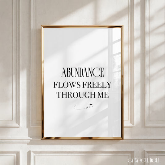 A framed typographic wall art print with the text 'ABUNDANCE FLOWS FREELY THROUGH ME' in black serif lettering on a white background, set in a thin gold frame and displayed against a white panelled wall with soft window shadows.