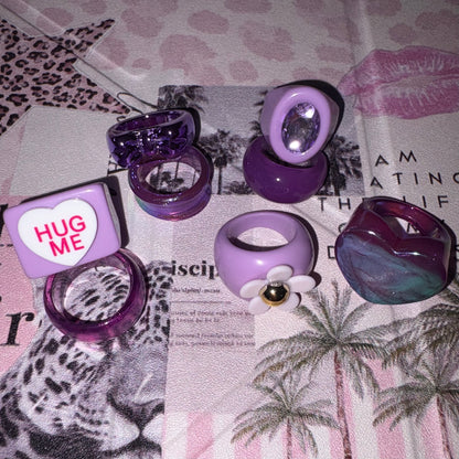 A collection of purple and lavender acrylic rings and a matching cube charm displayed on a pink and white palm tree patterned surface. The set includes a heart-shaped ring, a flower ring with a gold centre, a cube charm with a 'HUG ME' heart, and several translucent purple rings.
