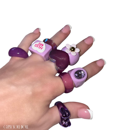 A close-up view of a hand with French-tip manicures crossing over each other, modelling a chunky pastel purple acrylic candy ring stack by Gypsy Soul Doll that features a matte lilac 3D flower ring, a square 'HUG ME' conversation heart ring, a glossy translucent plum heart ring, and chunky resin bands with embedded violet rhinestones against a clean white background.