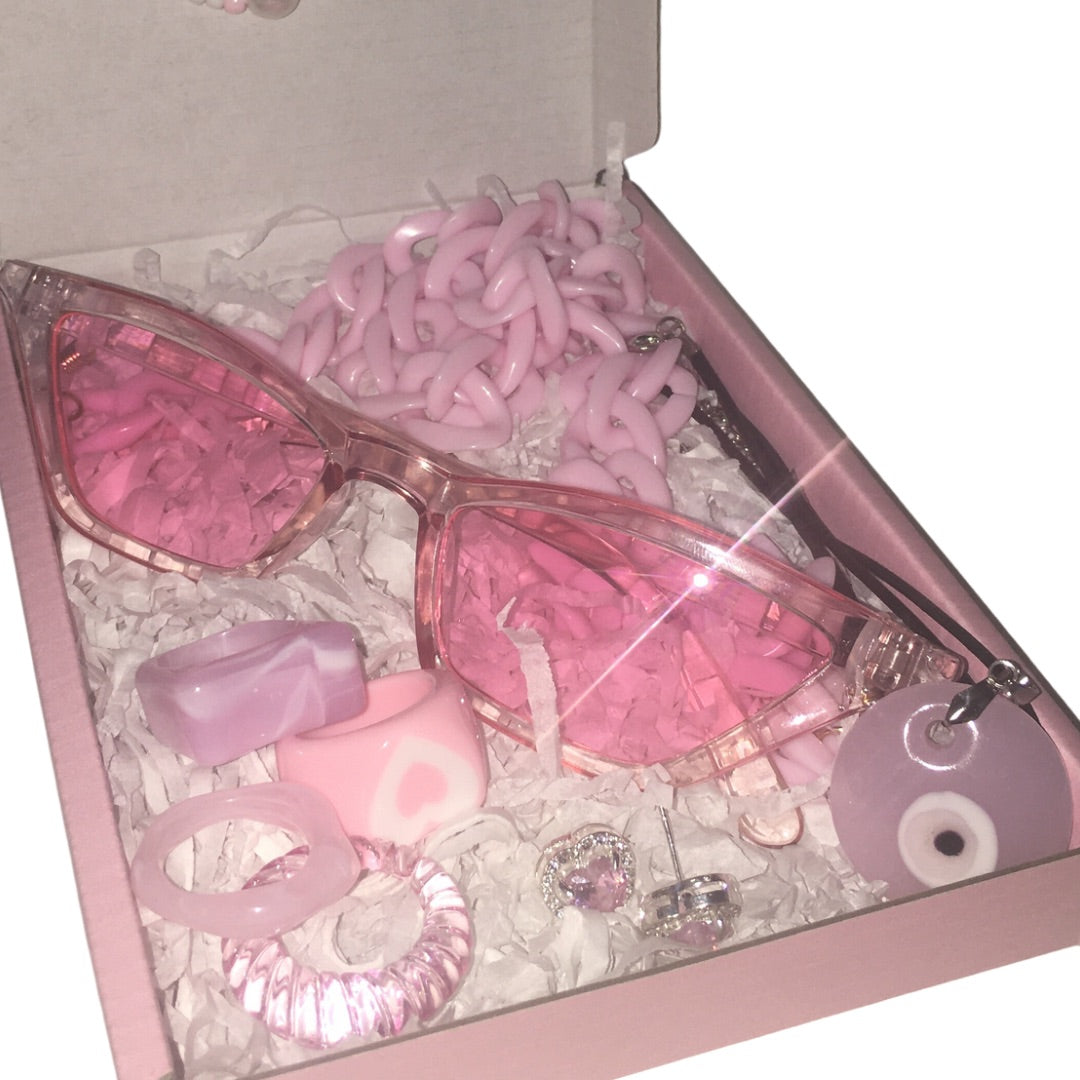 A premium pink boutique gift box filled with white crinkle paper shredded packaging, displaying a pair of translucent bubblegum pink cat-eye fashion glasses with a chunky pink chain lanyard strap, a set of pink and lavender marbled acrylic rings including a heart design, pink rhinestone heart stud earrings, and a matching pink evil eye charm pendant.